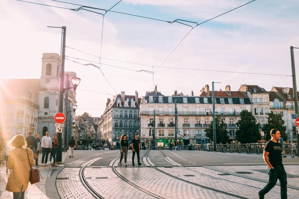 People walking across a sunny tramway intersection in a historic European city.